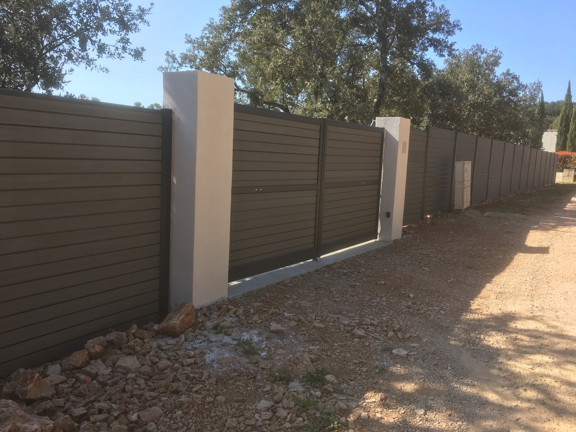 Clôture composite et portail moderne avec piliers blancs Portail et clôture en bois composite gris avec piliers blancs modernes devant une forêt sous un ciel bleu.