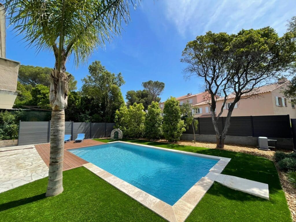 Piscine rectangulaire entourée de gazon synthétique, terrasse en bois, palmier et transats sous un ciel bleu ensoleillé.