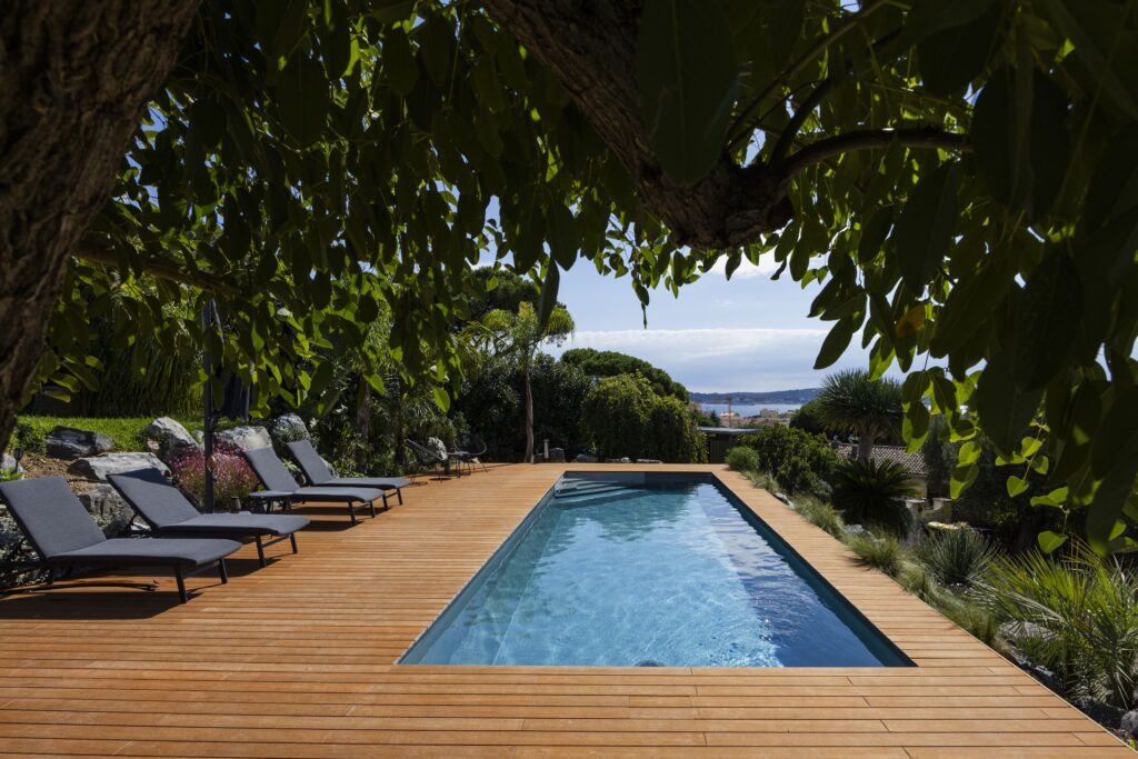 Piscine avec terrasse en bois, transats et vue mer dans un jardin méditerranéen luxuriant.