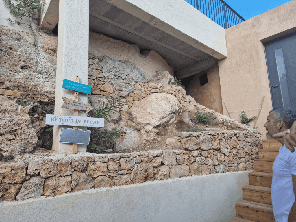 Panneaux déco 'Retour de pêche' et 'Agay' sur mur de pierre. Homme pointant des écriteaux en bois et pastis pétanque.